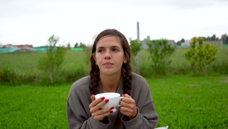 Young-peaceful-woman-sits-on-grass-lawn-and-drinks-tea-from-cup,-frontal