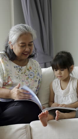 Senioren-Lifestyle-Konzept:-Eine-Liebevolle-Asiatische-Großmutter-Bringt-Ihrer-Enkelin-Geduldig-Das-Lesen-Und-Schreiben-Bei,-Während-Sie-Im-Gemütlichen-Komfort-Ihres-Wohnzimmers-Auf-Einem-Sofa-Sitzt