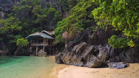 Deserted-Tropical-Island,-Rustic-Wooden-House-Under-Limestone-Cliffs,-Beach,-Trees-and-Turquoise-Sea