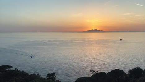 Zeitraffer-Des-Sonnenuntergangs-über-Den-Atemberaubenden-Küstenklippen-Von-Sorrento-In-Der-Nähe-Der-Amalfiküste,-Italien,-Während-Die-Feurige-Sonne-Hinter-Den-Bergen-Einer-Großen-Insel-Versinkt-Und-Den-Himmel-In-Leuchtende-Orangetöne-Taucht