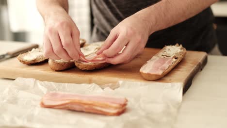 Kaukasischer-Mann-Legt-In-Der-Küche-Vorsichtig-Speckstreifen-Auf-Geschnittenes-Baguette