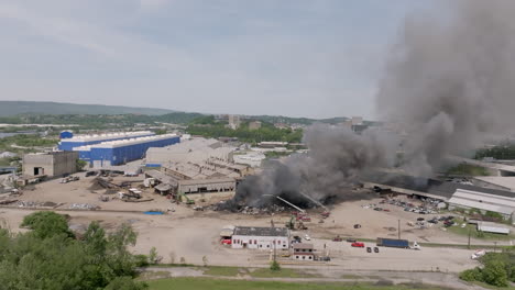 Wide-aerial-shot-rotating-around-a-large-scrap-metal-fire-with-emergency-services-covering-it-with-water-as-black-smoke-moves-into-the-sky