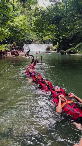 People-wearing-safety-jackets-and-float-in-a-row-on-the-mountain-river-when-on-vacation-together-with-waterfall-on-the-background