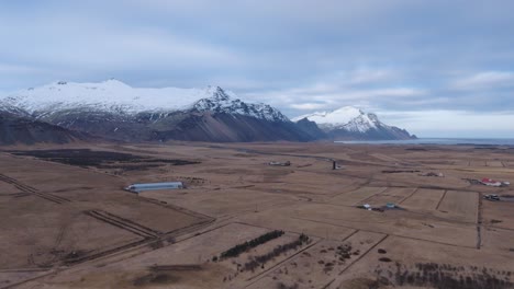 Fields-and-farm-buildings-in-Iceland-countryside-below-snowy-mountains