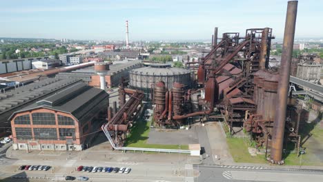 Aerial-view-of-an-old,-large,-rusted-industrial-complex-surrounded-by-roads-and-overgrown-greenery-under-a-clear-sky