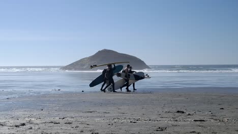 Un-Grupo-De-Surfistas-Camina-Con-Tablas-De-Surf-En-La-Soleada-Pista-De-Playa-Slomo.
