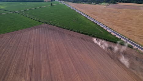 Tractor-ploughs-dirt-field-to-angled-edge-with-dust-cloud-rising-behind-in-lush-field