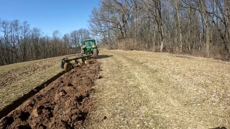 Siguiendo-Un-Tractor-John-Deere-Remolcando-Un-Arado-A-Través-De-Un-Campo-Inactivo-A-Principios-De-La-Primavera-En-El-Medio-Oeste