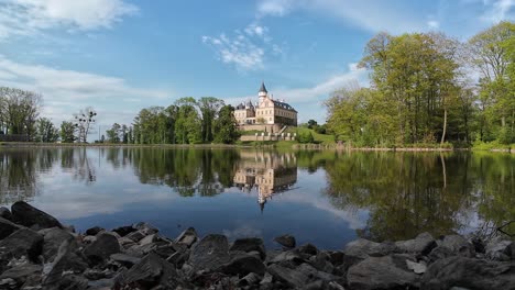 Majestic-castle-mirrored-in-the-tranquil-waters-of-a-lake,-nestled-amidst-lush-greenery-under-a-clear,-blue-sky