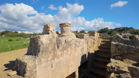 Partially-standing-stone-structure-at-Kato-Pafos-with-a-single-column-base-on-top,-set-against-a-bright-blue-sky-and-fluffy-clouds