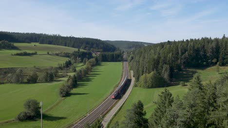A-train-traveling-through-a-lush,-green-valley-flanked-by-dense-forests,-aerial-view