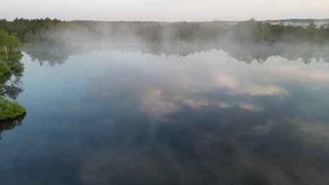 Morning-fog-rises-off-reflective-surface-of-northern-lake,-flyover