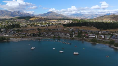 Wanaka-lakefront,-boats-moored-in-blue-water-against-a-backdrop-of-mountains-and-town,-aerial-view