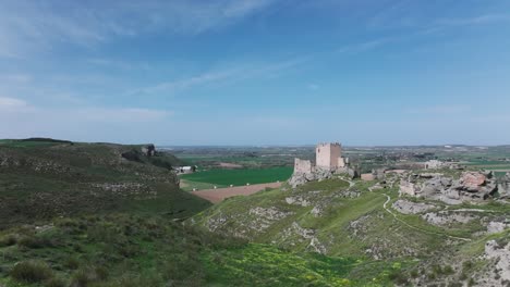 drone-flight-taking-off-from-a-platform-viewing-the-9th-century-Oreja-castle-and-the-remains-of-its-walls-behind-in-an-intense-green-there-are-crop-fields-with-a-blue-sky-in-Ontigola-Toledo-Spain