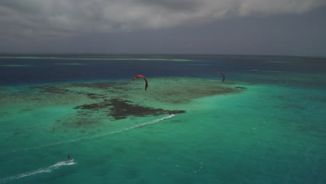 Two-kite-surfers-gliding-over-a-vibrant-coral-reef-in-clear-turquoise-waters,-aerial-view