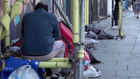 Un-Hombre-Sin-Hogar-Está-Sentado-Fumando-Un-Cigarrillo-Junto-A-Una-Segunda-Persona-Que-Yace-En-Una-Tienda-De-Campaña-Roja-Rodeada-De-Pertenencias-De-La-Gente-Mientras-Miembros-Del-Público-Pasan.