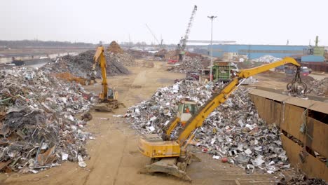 Gelbe-Bagger-Sortieren-Durch-Riesige-Haufen-Metallschrott-Auf-Einem-Industriellen-Recyclinghof