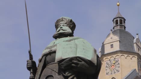 Statue-of-a-bearded-knight-holding-a-spear,-with-a-historic-clock-tower-in-the-background,-under-a-clear-sky