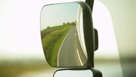 Rear-view-mirror-reflecting-a-sunny-rural-road-with-cars-in-the-distance,-blurred-vehicle-edge