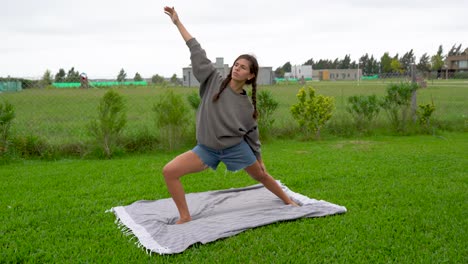 Una-Mujer-Joven-Hace-Posturas-De-Yoga-Guerrera-Sobre-Césped-En-Un-Día-Nublado-Y-Ventoso