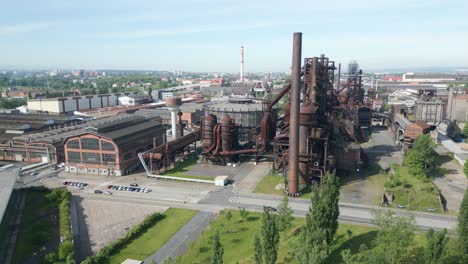 Aerial-view-of-an-old,-industrial-plant-with-large-rusted-metal-structures,-surrounded-by-greenery-and-a-road,-under-a-clear-sky