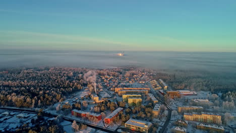 Aerial-View-of-Small-Industrial-Town-on-Sunny-Winter-Day,-Snow-Capped-Buildings-and-Forest-WIth-Coal-Power-Plant-in-Misty-Horizon