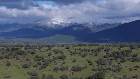 Vuelo-Lateral-Con-Dron-Visualizando-Una-Ladera-Con-Robles-Y-Detrás-Las-Montañas-De-Gredos-Con-Picos-Nevados-Y-Nubes-Ganchudas-Creando-Un-Efecto-De-Paralaje-Valle-Del-Tiétar-ávila-España