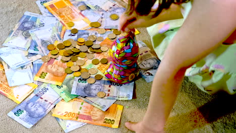 Girl-takes-coins-on-top-of-South-African-banknotes-and-deposits-into-money-box