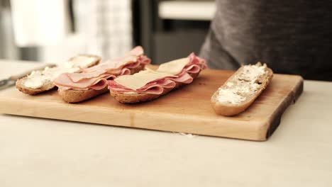 Hombre-Caucásico-Colocando-Queso-Suizo-En-Rodajas-En-Una-Baguette-Marrón-Cortada-A-La-Mitad-En-La-Cocina-Sobre-La-Encimera