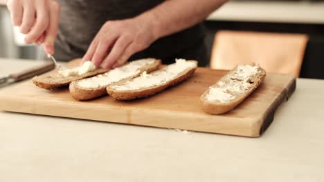 Kaukasischer-Mann,-Der-Cremigen-Käse-Auf-Baguette-Aus-Braunem-Mehl-Auf-Der-Küchenarbeitsplatte-Verteilt