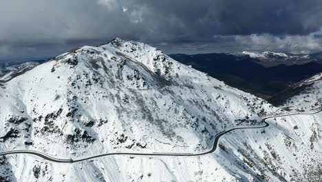 A-snow-covered-mountain-with-a-winding-road,-under-cloudy-skies,-aerial-view