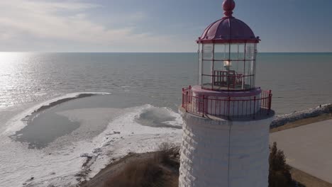 A-snow-covered-lighthouse-by-a-frozen-lake-under-clear-skies,-aerial-view