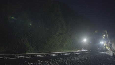 Nachtaufnahme-Von-Eisenbahnwartungsmaschinen-Bei-Der-Arbeit,-Beleuchtet-Durch-Helle-Lichter,-Umgeben-Von-Dichtem-Laub