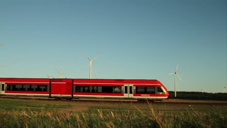 Red-train-crossing-rural-landscape-with-wind-turbines-against-a-clear-blue-sky,-daytime