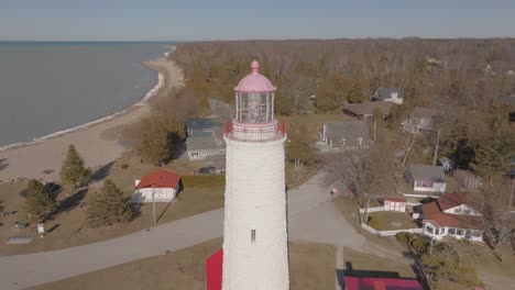 A-white-lighthouse-with-a-red-top-near-a-wooded-coastal-area-on-a-clear-day,-aerial-view