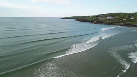 Flying-drone-backwards-over-waves-and-surfers,-revealing-more-of-the-landscape-in-West-Cork-Ireland
