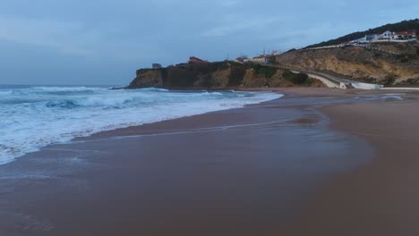 Low-drone-shot-of-waves-crushing-on-sand-Magoito-Beach-in-Portugal,-cloudy-sunset