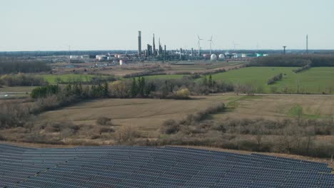 Wide-view-of-solar-panels-with-industrial-backdrop-and-wind-turbines,-vibrant-green-fields-surround-the-area,-daytime,-sunny