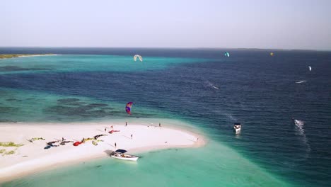 Kite-surfers-at-punta-madrisky-with-vibrant-turquoise-waters-and-busy-beach-activities,-aerial-view