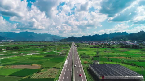 Beautiful-Peaceful-Highway-By-Farmland-Fields-With-Electricity-Pylons-And-Car-Traffic-Cargo-Truck-Transportation-Under-Blue-Sky-Clouds-Forward-To-Mountains