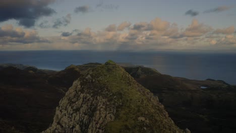 Aerial-atop-a-rocky-peak-in-the-Scottish-Highlands-reveals-the-ocean-and-cloudy-sky-on-the-horizon,-Isle-of-Skye,-Scotland