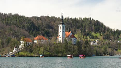 Kirche-Am-Bleder-See-Und-Traditionelle-Pletna-Boote-Mit-Touristen