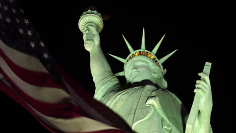 Bandera-Americana-Ondeando-Bajo-La-Estatua-De-La-Libertad-Por-La-Noche,-Cerrar-En-ángulo-Bajo