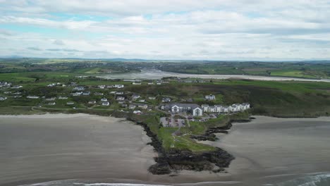 Flying-backwards-and-revealing-panorama-over-Inchidoney-beach-with-cliffs,-hill-and-village-in-West-Cork,-Ireland