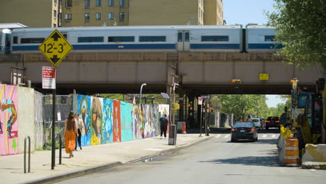 People-Walk-on-Harlem-Street-Under-Elevated-Train-on-Sunny-Day,-New-York-City