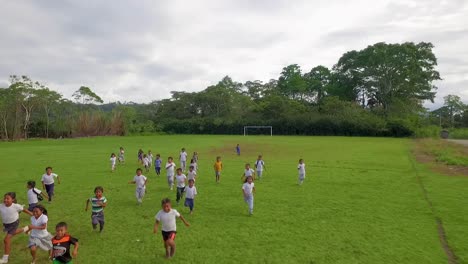 Group-of-laughing-school-children-running-towards-the-camera-in-a-green-football-field-playing-in-uniform-in-jungle-and-forest-of-Ecuador---Drone-reveal-shot