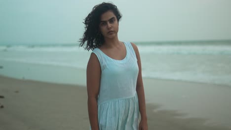 Woman-in-white-dress-stands-contemplatively-on-a-cloudy-beach-at-dusk,-waves-gently-breaking