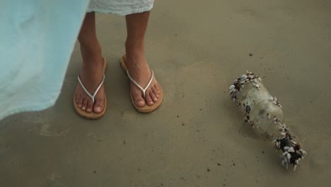 Woman-in-flip-flops-discovers-a-message-bottle-covered-in-shells-on-sandy-beach