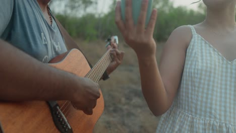 Nahaufnahme-Eines-Mannes,-Der-Neben-Einer-Frau-Im-Sommerkleid-Gitarre-Spielt,-Im-Freien-In-Der-Abenddämmerung