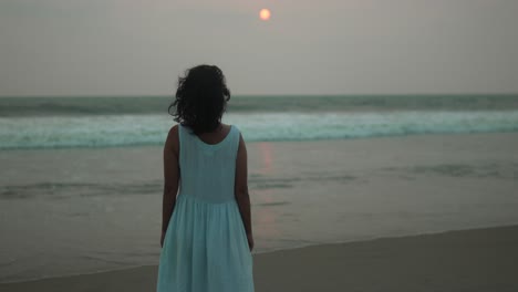 Young-girl-in-white-dress-standing-on-beach-at-sunset,-watching-waves